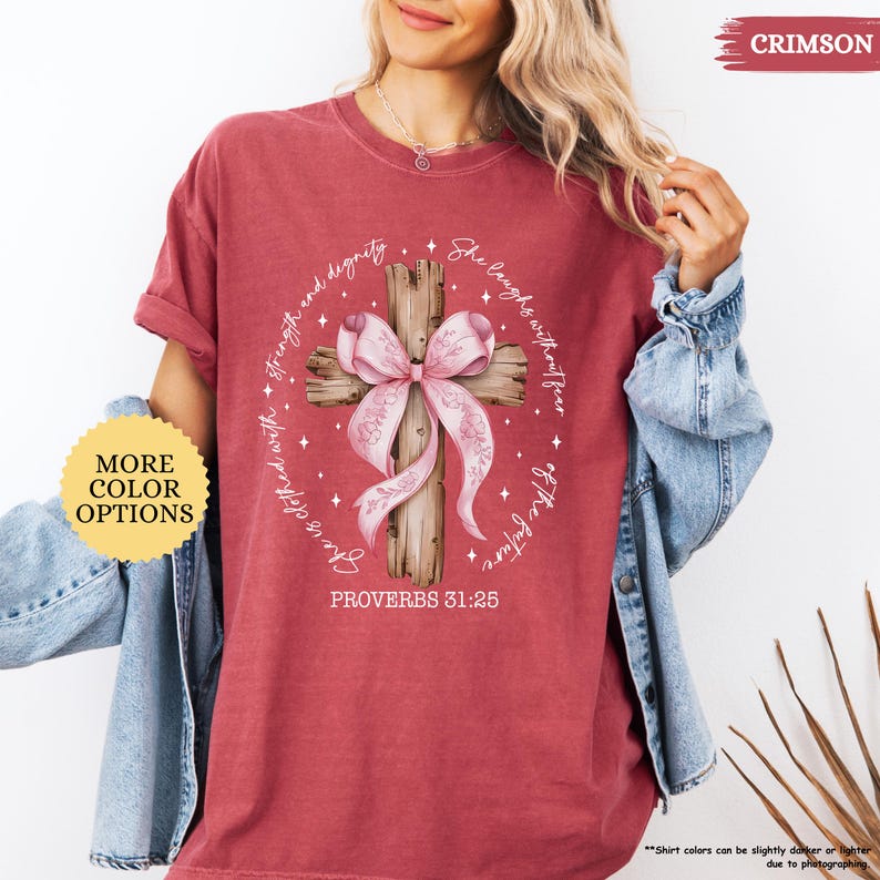 Op de afbeelding: Een roodbruin t-shirt met een verweerde look heeft een houten kruis met een roze strik. Het kruis is omringd door een cirkel van witte sterren en de tekst "Zij lacht zonder angst voor de toekomst" en "Zij is bekleed met kracht en waardigheid". De onderkant van het ontwerp bevat de tekst "Spreuken 31:25".