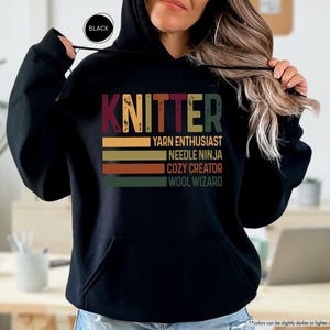 May include: Black hooded sweatshirt with the word "KNITTER" in colorful letters. Below the word are the phrases "YARN ENTHUSIAST", "NEEDLE NINJA", "COZY CREATOR", and "WOOL WIZARD" in a retro style.