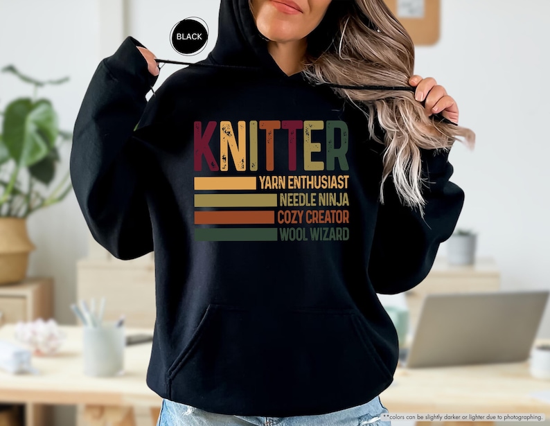 May include: Black hooded sweatshirt with the word "KNITTER" in colorful letters. Below the word are the phrases "YARN ENTHUSIAST", "NEEDLE NINJA", "COZY CREATOR", and "WOOL WIZARD" in a retro style.