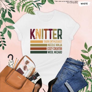 May include: White t-shirt with the text "KNITTER" in a vintage style font. The text is layered over colorful stripes with the words "Yarn Enthusiast", "Needle Ninja", "Cozy Creator", and "Wool Wizard" underneath.