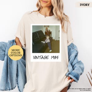 Op de afbeelding: Een beige t-shirt met een vintage foto van een jonge jongen die op een bank zit. De foto is omlijst met een witte rand en de tekst "Vintage 1985" is onder de foto gedrukt.
