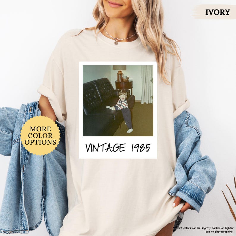 Op de afbeelding: Een beige t-shirt met een vintage foto van een jonge jongen die op een bank zit. De foto is omlijst met een witte rand en de tekst "Vintage 1985" is onder de foto gedrukt.