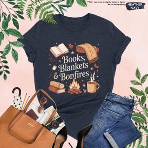 May include: Heather navy t-shirt with the text "Books, Blankets & Bonfires" in a whimsical design. The graphic includes a book, blanket, campfire, and mug. The shirt is paired with blue jeans and a brown tote bag.