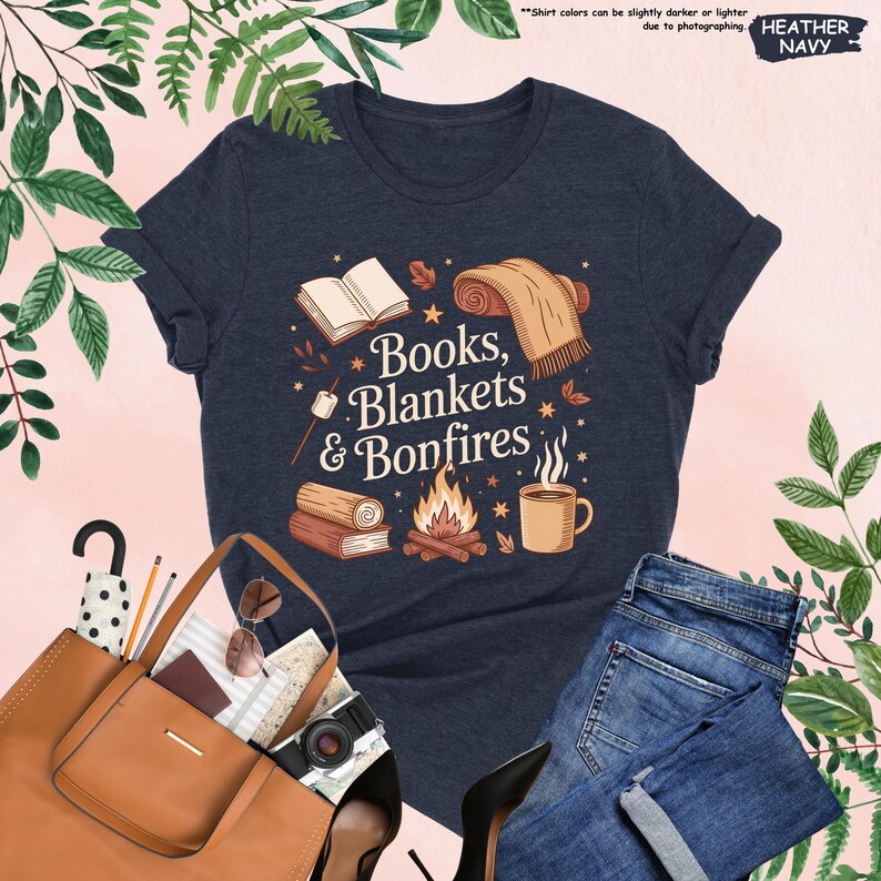 May include: Heather navy t-shirt with the text "Books, Blankets & Bonfires" in a whimsical design. The graphic includes a book, blanket, campfire, and mug. The shirt is paired with blue jeans and a brown tote bag.