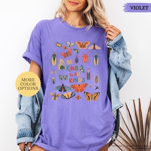 May include: A lilac t-shirt with a design of various insects and butterflies. The text "BE KIND TO ALL KINDS" is printed on the front. A button on the left says "MORE COLOR OPTIONS".