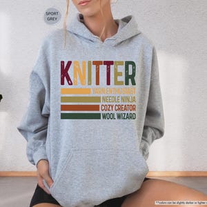 May include: Sport grey hoodie with a front graphic that reads "KNITTER" in maroon, green, and gold. Below the word "KNITTER" are the phrases "YARN ENTHUSIAST", "NEEDLE NINJA", "COZY CREATOR", and "WOOL WIZARD".