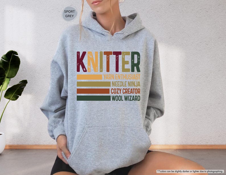 May include: Sport grey hoodie with a front graphic that reads "KNITTER" in maroon, green, and gold. Below the word "KNITTER" are the phrases "YARN ENTHUSIAST", "NEEDLE NINJA", "COZY CREATOR", and "WOOL WIZARD".