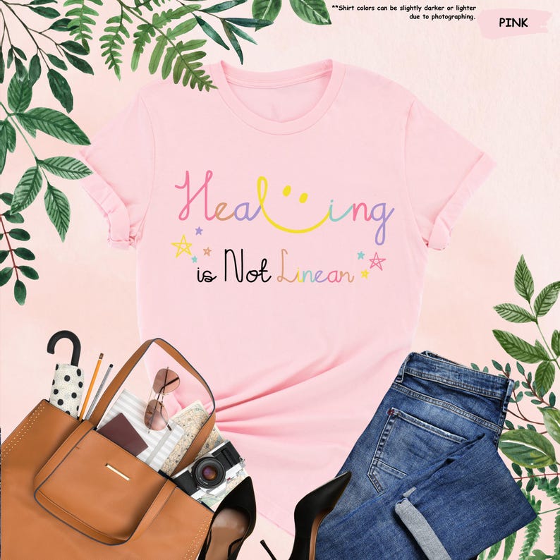 K&ouml;nnte beinhalten: Rosa T-Shirt mit dem Text "Healing is Not Linear" in bunten Buchstaben mit einem Smiley und Sternen.