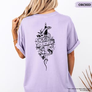 May include: A lilac-coloured t-shirt with a black graphic of a snake and dagger design, with the words "you do not yield" on a banner. The design is surrounded by floral elements. The shirt's colour is a light purple.