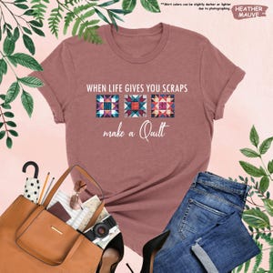 May include: Heather mauve t-shirt with the text "WHEN LIFE GIVES YOU SCRAPS make a Quilt." The shirt features three colorful quilt block designs. The shirt is paired with blue jeans, a brown purse, and black heels. The words "HEATHER MAUVE" are also visible.