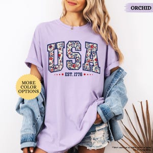 May include: A purple t-shirt with a floral print design that spells out "USA" with the text "EST. 1776" underneath. The shirt is being modeled by a person wearing denim shorts and a denim jacket.