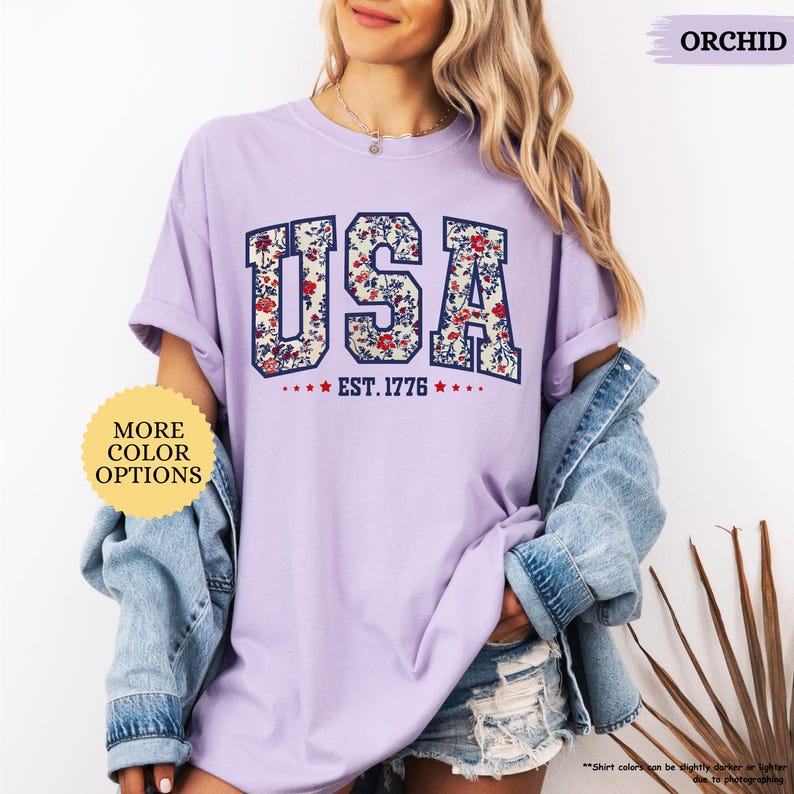 May include: A purple t-shirt with a floral print design that spells out "USA" with the text "EST. 1776" underneath. The shirt is being modeled by a person wearing denim shorts and a denim jacket.
