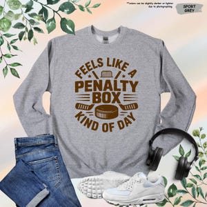May include: Sport grey sweatshirt with the text "Feels Like A Penalty Box Kind of Day" in brown. The design includes hockey sticks, a puck, and a net. The sweatshirt is paired with blue jeans and white sneakers.
