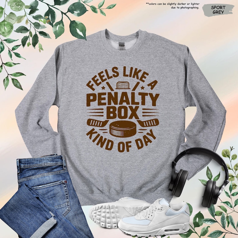 May include: Sport grey sweatshirt with the text "Feels Like A Penalty Box Kind of Day" in brown. The design includes hockey sticks, a puck, and a net. The sweatshirt is paired with blue jeans and white sneakers.