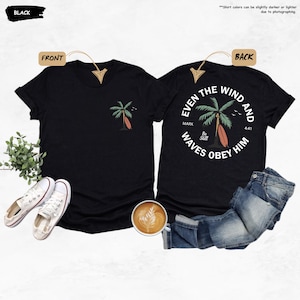 K&ouml;nnte beinhalten: Schwarze T-Shirts mit einem Palmen- und Surfbrett-Motiv. Die Vorderseite des Shirts hat ein kleines Design, w&auml;hrend die R&uuml;ckseite ein kreisf&ouml;rmiges Design mit dem Text "Even the wind and waves obey him" aufweist.