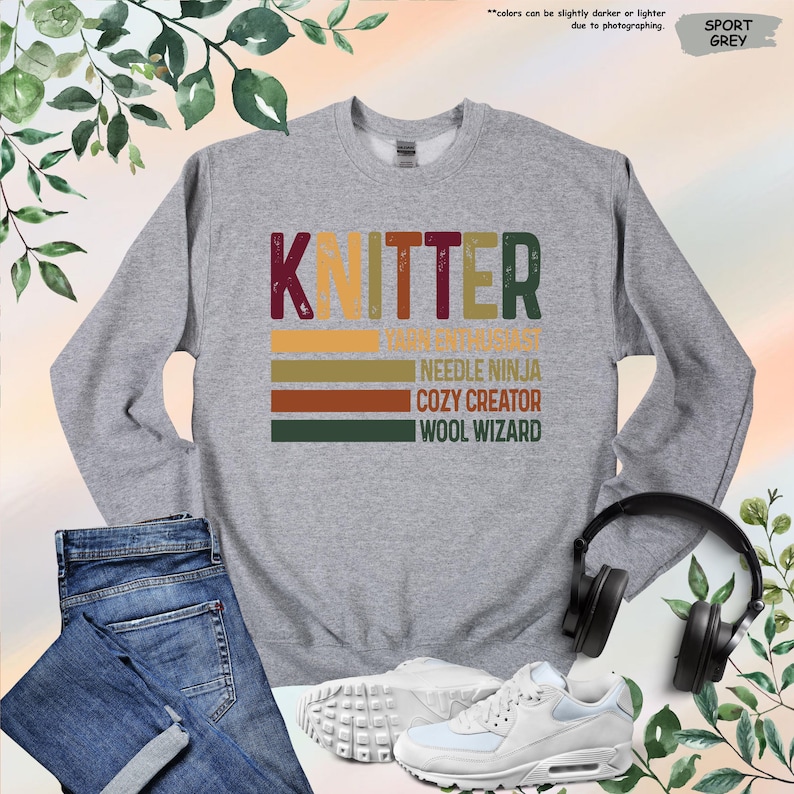 May include: A heather grey sweatshirt with the word "KNITTER" in a distressed, colorful font. Below the word are the phrases "NEEDLE NINJA", "COZY CREATOR", and "WOOL WIZARD". The sweatshirt is paired with blue jeans and white sneakers.