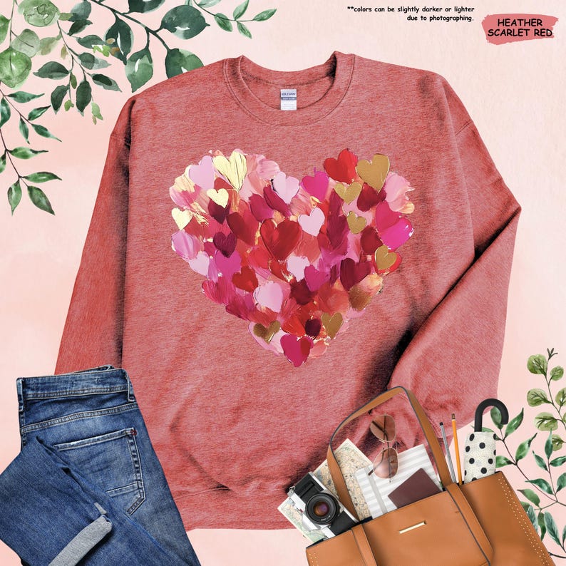 Pu&ograve; includere: Felpa rosso scarlatto heather con un design a cuore realizzato con pennellate stratificate nei toni del rosso, rosa e oro. Il testo "colors can be slightly darker or lighter due to photographing" &egrave; visibile.