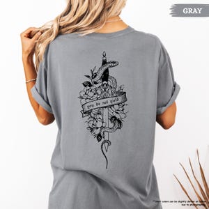 May include: A grey t-shirt with a black graphic on the back. The graphic depicts a snake entwined around a sword, with flowers and the text "you do not yield". The shirt is a solid grey colour.
