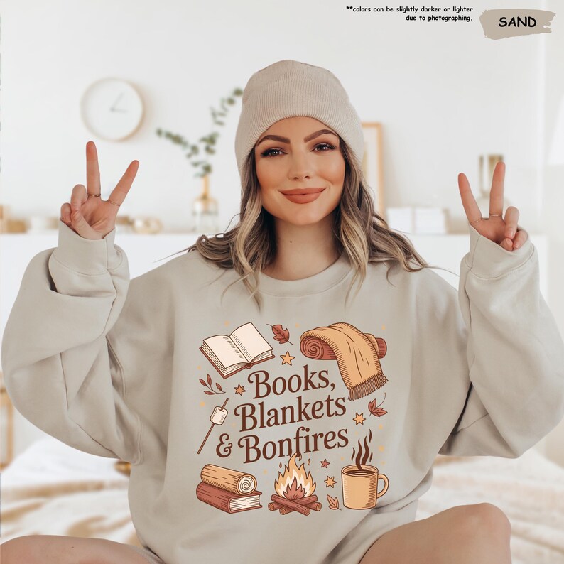 May include: Sand-colored sweatshirt with the text "Books, Blankets & Bonfires" and illustrations of a book, blanket, campfire, and mug. The model is wearing a matching beanie. The text "colors can be slightly darker or lighter due to photographing" is also visible.