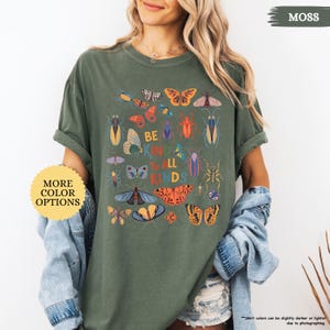 May include: A moss green t-shirt with a graphic of various insects and butterflies, and the text "BE KIND TO ALL KINDS." The shirt is worn with a denim jacket. A button on the left says "MORE COLOR OPTIONS." The shirt's sleeves are rolled up.