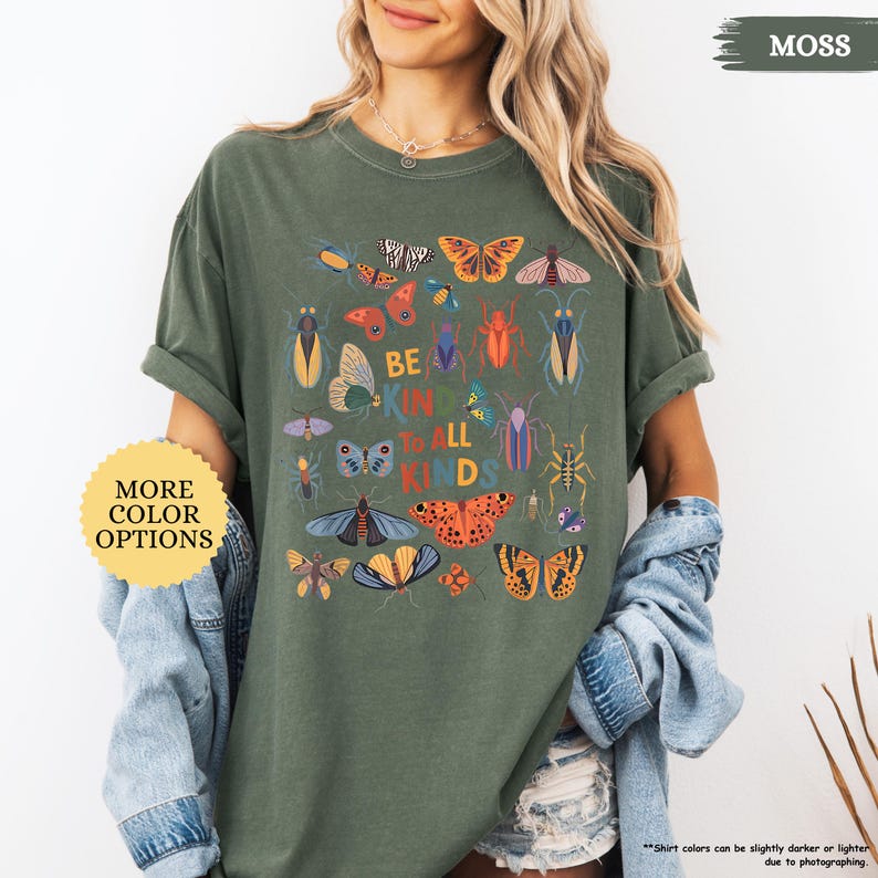 May include: A moss green t-shirt with a graphic of various insects and butterflies, and the text "BE KIND TO ALL KINDS." The shirt is worn with a denim jacket. A button on the left says "MORE COLOR OPTIONS." The shirt's sleeves are rolled up.