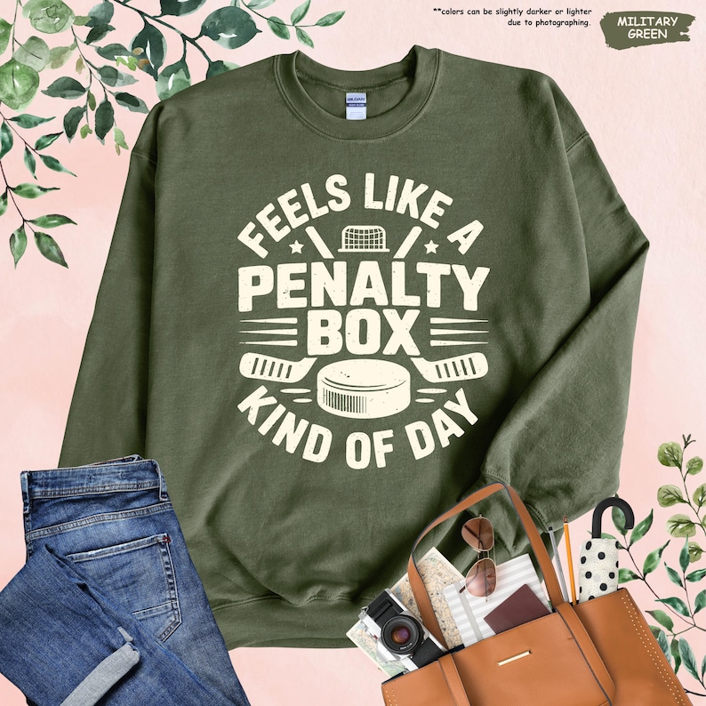 May include: An olive green sweatshirt with the text "FEELS LIKE A PENALTY BOX KIND OF DAY" in white. The design includes hockey sticks, a hockey puck, and a hockey net. The sweatshirt is paired with blue jeans and a brown tote bag.