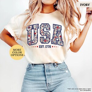 May include: A white t-shirt with a floral print design that spells out "USA" with the text "EST. 1776" underneath. The shirt is being modeled by a person wearing blue jeans.