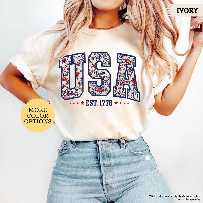 May include: A white t-shirt with a floral print design that spells out "USA" with the text "EST. 1776" underneath. The shirt is being modeled by a person wearing blue jeans.