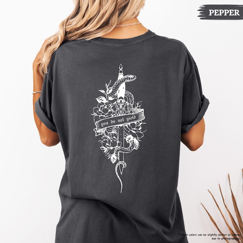 May include: Dark grey t-shirt with a white graphic on the back. The graphic includes a snake, sword, flowers, and the text "you do not yield." The shirt has a relaxed fit.