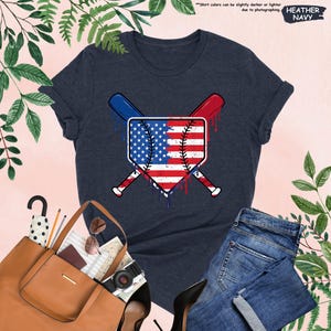 K&ouml;nnte beinhalten: Heather-Marineblaues T-Shirt mit einem Baseball-Diamanten-Design mit der amerikanischen Flagge und gekreuzten Schl&auml;gern. Das Design hat rote und blaue Akzente. Das Shirt wird mit einer blauen Jeans und einer braunen Tragetasche kombiniert.