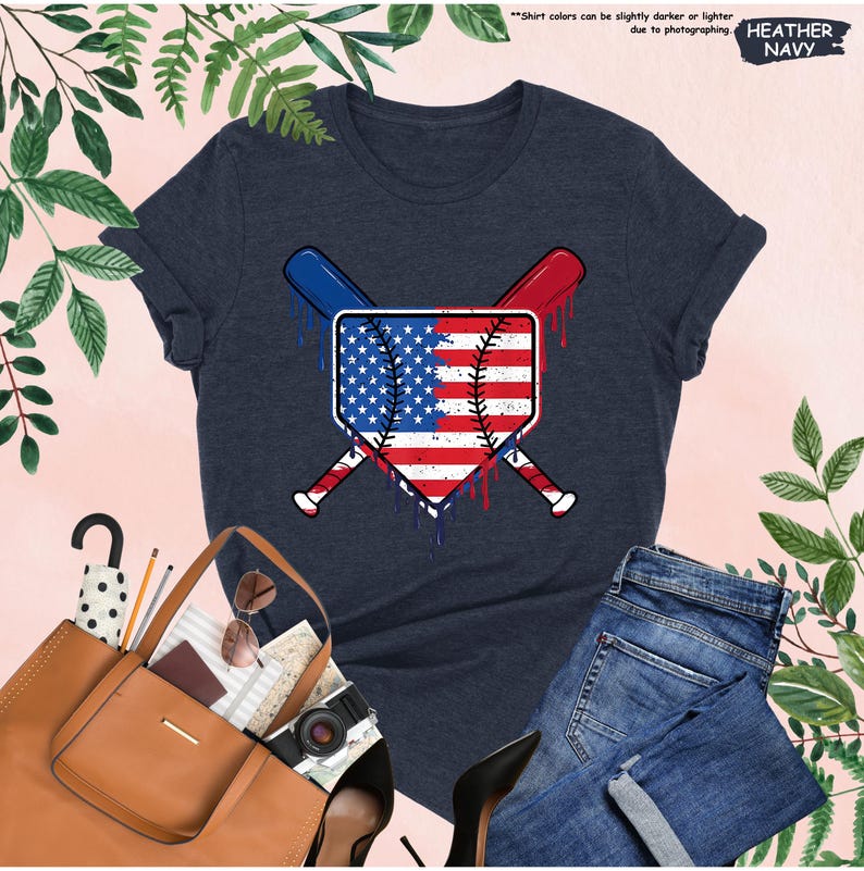 K&ouml;nnte beinhalten: Heather-Marineblaues T-Shirt mit einem Baseball-Diamanten-Design mit der amerikanischen Flagge und gekreuzten Schl&auml;gern. Das Design hat rote und blaue Akzente. Das Shirt wird mit einer blauen Jeans und einer braunen Tragetasche kombiniert.