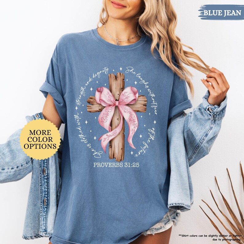 Op de afbeelding: Een jeansblauw T-shirt met een roze lint om een houten kruis gebonden. De tekst "She laughs without fear of the future" en "Proverbs 31:25" staat op het T-shirt gedrukt.