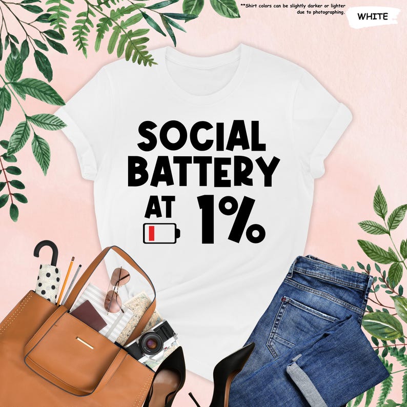 May include: White t-shirt with the text "SOCIAL BATTERY AT 1%" in black. The design includes a battery icon at 1%. The shirt is paired with blue jeans, a brown tote bag, and black heels.