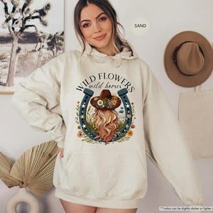 May include: A cream colored hoodie with a graphic of a woman wearing a cowboy hat and a horseshoe with the text "WILD FLOWERS wild horses" printed on the front.