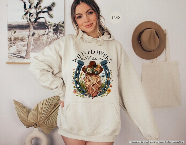 May include: A cream colored hoodie with a graphic of a woman wearing a cowboy hat and a horseshoe with the text "WILD FLOWERS wild horses" printed on the front.