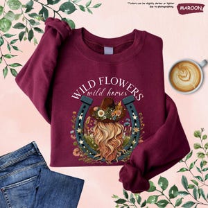 May include: Maroon sweatshirt with a graphic of a woman with long blonde hair wearing a cowboy hat and a horseshoe with flowers around it. The text "WILD FLOWERS wild horses" is printed on the sweatshirt.