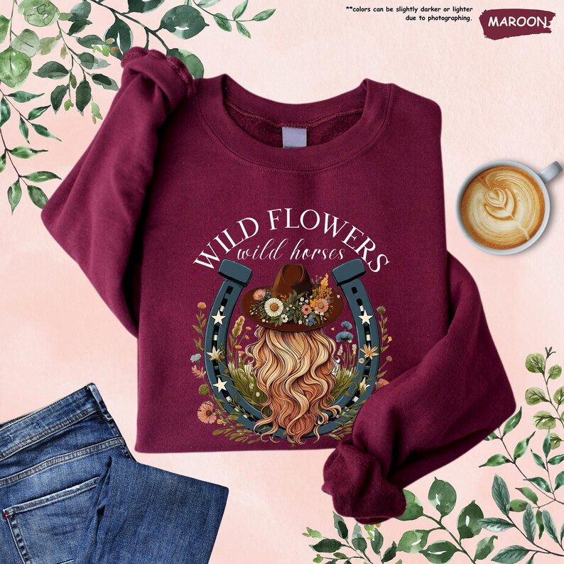 May include: Maroon sweatshirt with a graphic of a woman with long blonde hair wearing a cowboy hat and a horseshoe with flowers around it. The text "WILD FLOWERS wild horses" is printed on the sweatshirt.