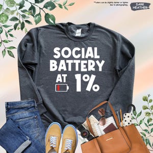 May include: Dark heather gray sweatshirt with the text "SOCIAL BATTERY AT 1%" in white. The design includes a battery icon at 1%. The image also shows blue jeans, tan shoes, a brown purse, and accessories.