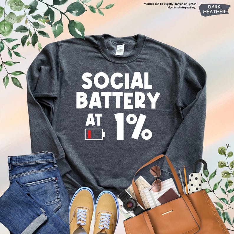 May include: Dark heather gray sweatshirt with the text "SOCIAL BATTERY AT 1%" in white. The design includes a battery icon at 1%. The image also shows blue jeans, tan shoes, a brown purse, and accessories.