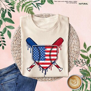 K&ouml;nnte beinhalten: Cremefarbenes T-Shirt mit einem Baseball-Homeplate-Design mit der amerikanischen Flagge und gekreuzten Baseballschl&auml;gern. Das Design verwendet die Farben Rot, Wei&szlig; und Blau mit einem tropfenden Farbeffekt. Das Shirt wird mit blauen Jeans und einer Kaffeetasse gezeigt.