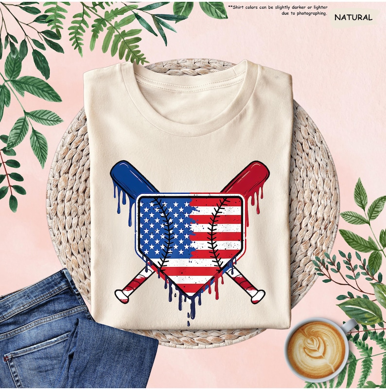 K&ouml;nnte beinhalten: Cremefarbenes T-Shirt mit einem Baseball-Homeplate-Design mit der amerikanischen Flagge und gekreuzten Baseballschl&auml;gern. Das Design verwendet die Farben Rot, Wei&szlig; und Blau mit einem tropfenden Farbeffekt. Das Shirt wird mit blauen Jeans und einer Kaffeetasse gezeigt.