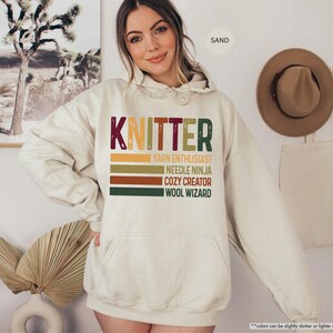 May include: A sand-colored hoodie with the word "KNITTER" in a retro font. Below the word are the phrases "YARN ENTHUSIAST", "NEEDLE NINJA", "COZY CREATOR", and "WOOL WIZARD" in a stacked design. The hoodie has a front pocket and a drawstring hood.