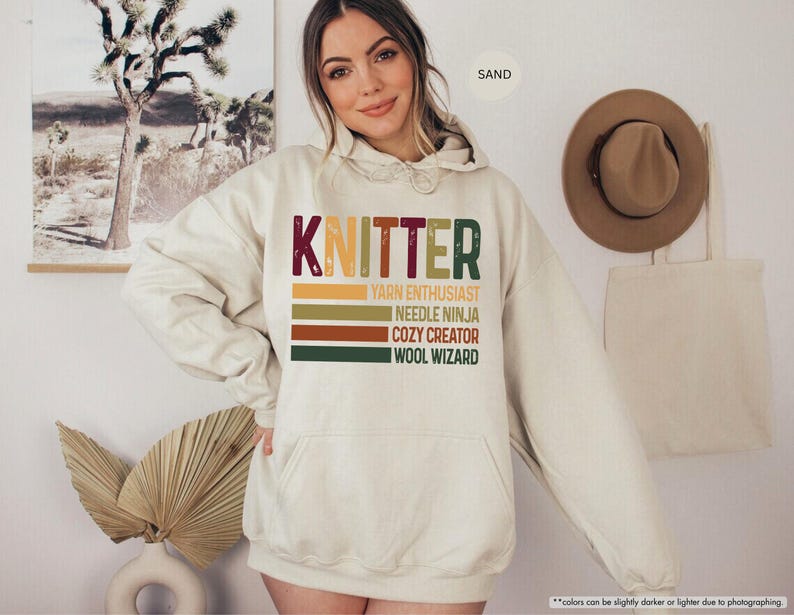 May include: A sand-colored hoodie with the word "KNITTER" in a retro font. Below the word are the phrases "YARN ENTHUSIAST", "NEEDLE NINJA", "COZY CREATOR", and "WOOL WIZARD" in a stacked design. The hoodie has a front pocket and a drawstring hood.