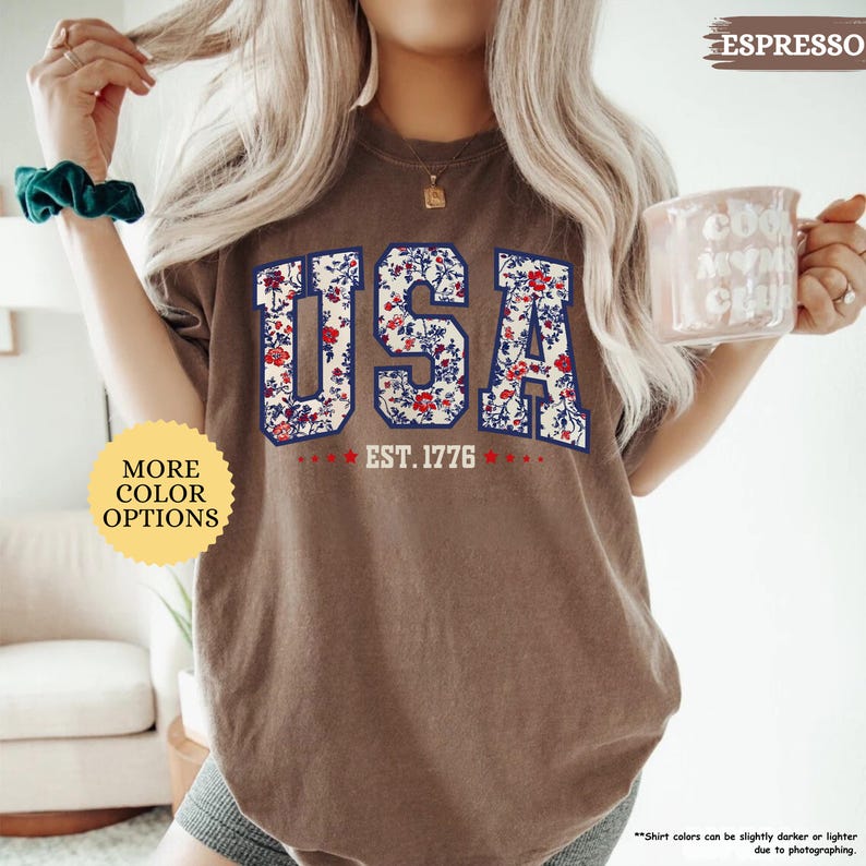 May include: A brown t-shirt with a floral print design that spells out "USA" with the text "EST. 1776" underneath. The shirt is being modeled by a person.
