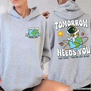 May include: Sport grey hoodie with a front pocket and a drawstring hood. The front features a graphic of a globe with the text "TOMORROW NEEDS YOU". The back displays an astronaut on a globe with the same text and "KEEP GOING - YOU ARE NOT ALONE".