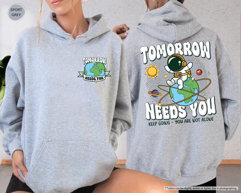 May include: Sport grey hoodie with a front pocket and a drawstring hood. The front features a graphic of a globe with the text "TOMORROW NEEDS YOU". The back displays an astronaut on a globe with the same text and "KEEP GOING - YOU ARE NOT ALONE".