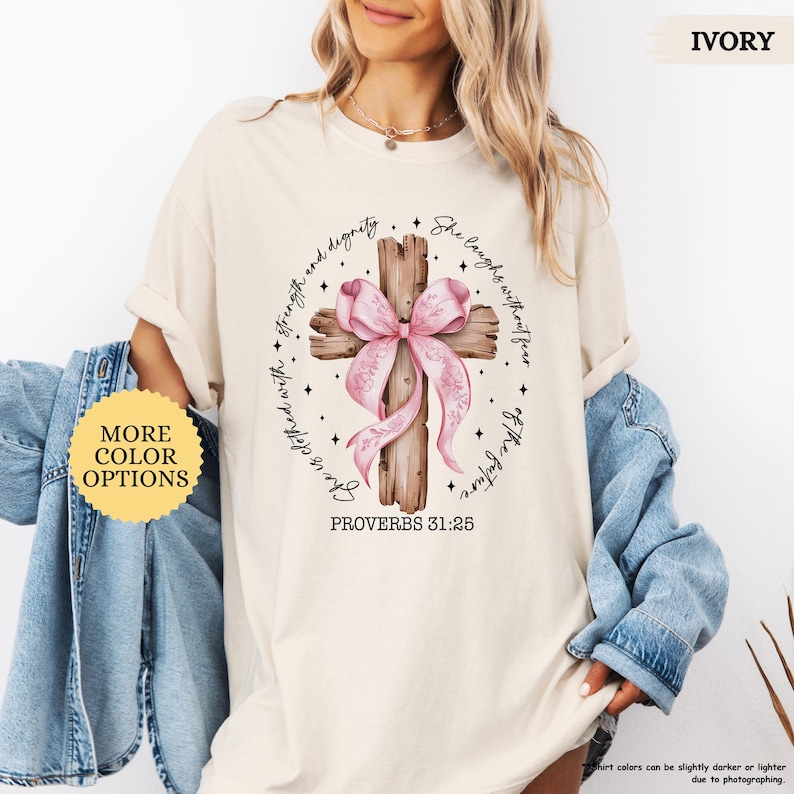 Op de afbeelding: Een beige T-shirt met een bruin houten kruis versierd met een roze lint en de tekst "Zij lacht zonder angst voor de toekomst" en "Spreuken 31:25".