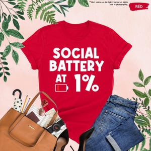 May include: A red t-shirt with the text "SOCIAL BATTERY AT 1%" in white. The shirt is paired with blue jeans, a brown tote bag, sunglasses, and a camera. The image includes greenery and the word "RED".