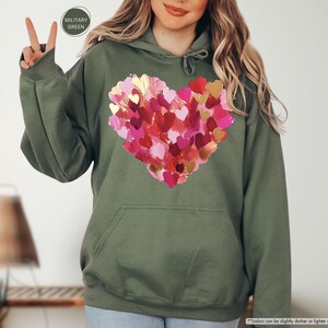 Pu&ograve; includere: Felpa con cappuccio verde oliva con un grande cuore composto da cuori rosa, rossi e dorati sovrapposti. La felpa con cappuccio ha un cappuccio con coulisse e una tasca frontale. Le parole "MILITARY GREEN" sono in un cerchio in alto a sinistra.