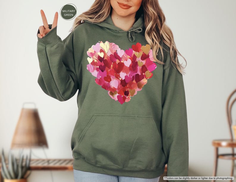 Pu&ograve; includere: Felpa con cappuccio verde oliva con un grande cuore composto da cuori rosa, rossi e dorati sovrapposti. La felpa con cappuccio ha un cappuccio con coulisse e una tasca frontale. Le parole "MILITARY GREEN" sono in un cerchio in alto a sinistra.
